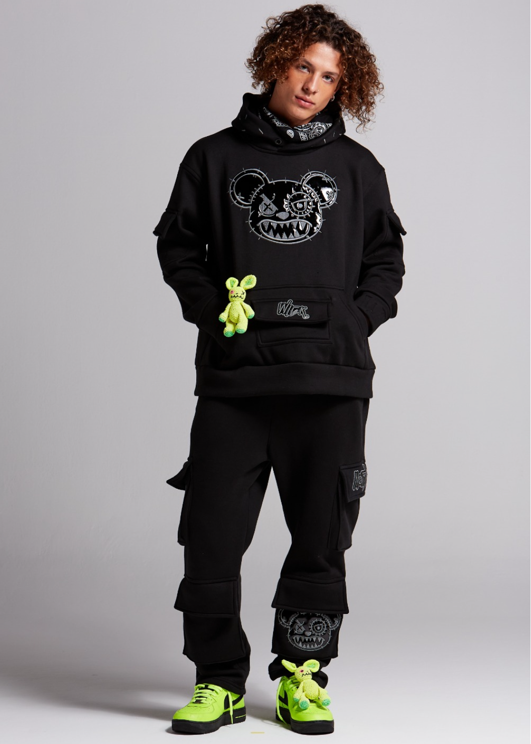 Person wearing a black hoodie with a graphic design, black cargo pants, and green sneakers on a gray background