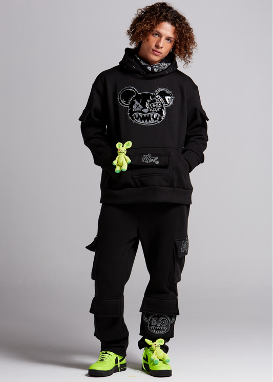 Person wearing a black hoodie with a graphic design, black cargo pants, and green sneakers on a gray background