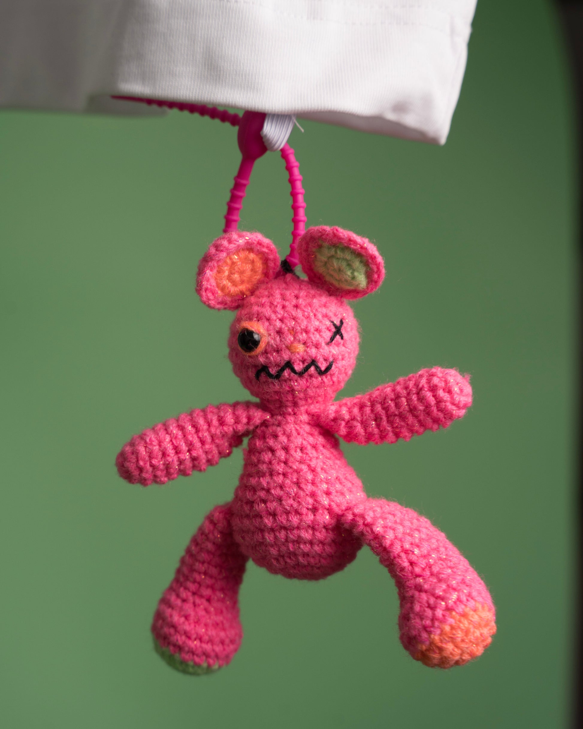 Pink crocheted creature with a sad face hanging from a string against a green background