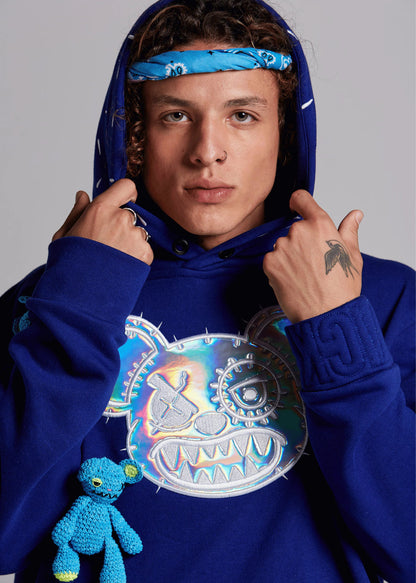 Person wearing a blue hoodie with a colorful graphic design, holding a small blue plush toy.
