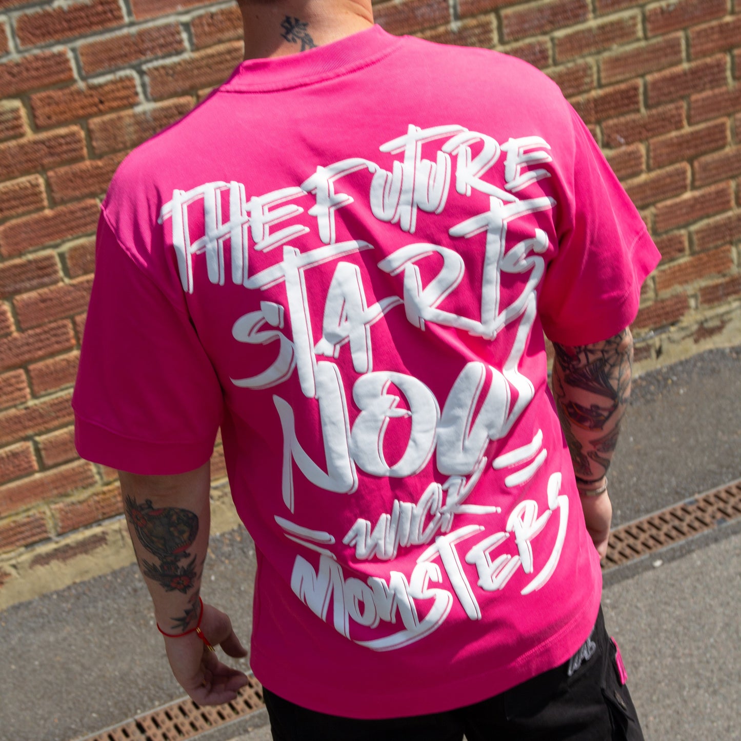 Person wearing a pink t-shirt with white text on the back, standing against a brick wall.